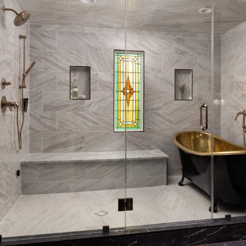 Luxurious wet room featuring marble-look tile walls and floors, a walk-in shower with built-in bench, dual showerheads, and recessed niches. A freestanding black clawfoot tub with a gold interior sits beneath a stained glass window, creating a spa-like, elegant atmosphere.