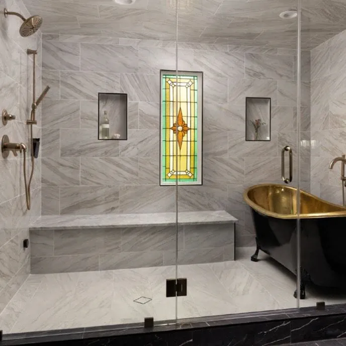 Luxurious wet room featuring marble-look tile walls and floors, a walk-in shower with built-in bench, dual showerheads, and recessed niches. A freestanding black clawfoot tub with a gold interior sits beneath a stained glass window, creating a spa-like, elegant atmosphere.