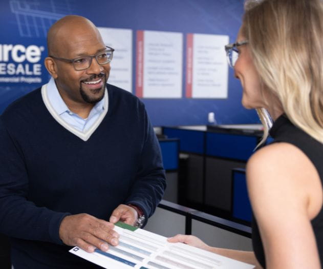 Two individuals interacting at a counter in a professional setting with signage in the background.
